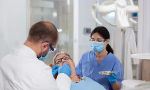 Dentysta zakłada białe plomby na NFZ starszej pacjentce. Asystentka stomatologiczna pomaga.