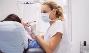 Dentystka w masce i rękawiczkach bada pacjentkę, która leży na fotelu. Może to być kontrola po zabiegu, np. po zerwanych szwach na dziąśle.