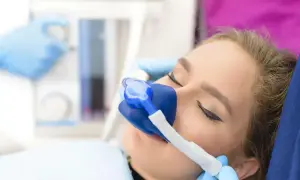 Pacjentka w trakcie leczenia w sedacji, z maską tlenową na twarzy. Dłonie w niebieskich rękawiczkach przytrzymują maskę.