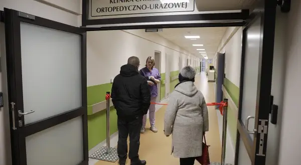 Korytarz szpitalny z tabliczką "Klinika Chirurgii Ortopedyczno-Urazowej", pacjenci i lekarka.
