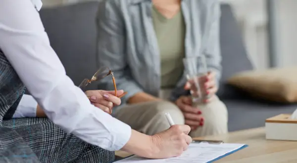 Psychoterapeutka pisze na dokumentach, druga osoba trzyma szklankę wody.
