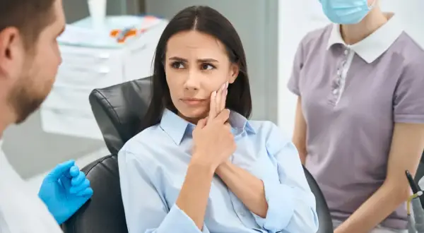 Pacjentka z bólem zęba, wskazująca na policzek, martwiąca się powikłaniami po szlifowaniu zębów. Obok dentysta i asystentka.