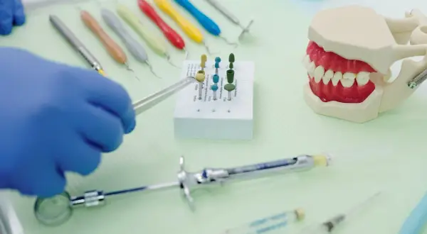 Rękawiczka w niebieskiej rękawiczce trzyma narzędzie do czyszczenia protezy zębowej sodą. Obok model szczęki i narzędzia dentystyczne.