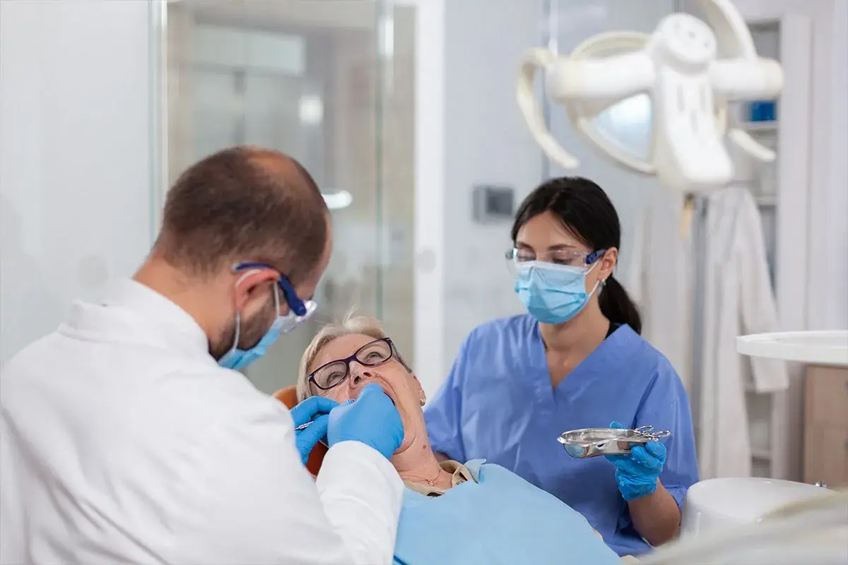 Dentysta zakłada białe plomby na NFZ starszej pacjentce. Asystentka stomatologiczna pomaga.