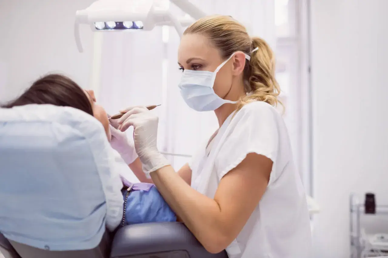 Dentystka w masce i rękawiczkach bada pacjentkę, która leży na fotelu. Może to być kontrola po zabiegu, np. po zerwanych szwach na dziąśle.