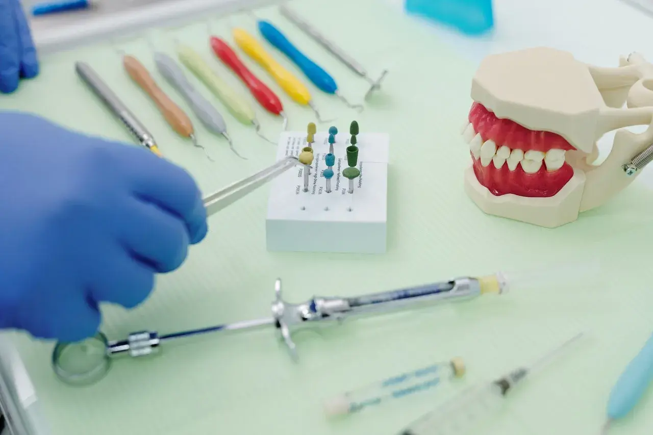 Rękawiczka w niebieskiej rękawiczce trzyma narzędzie do czyszczenia protezy zębowej sodą. Obok model szczęki i narzędzia dentystyczne.