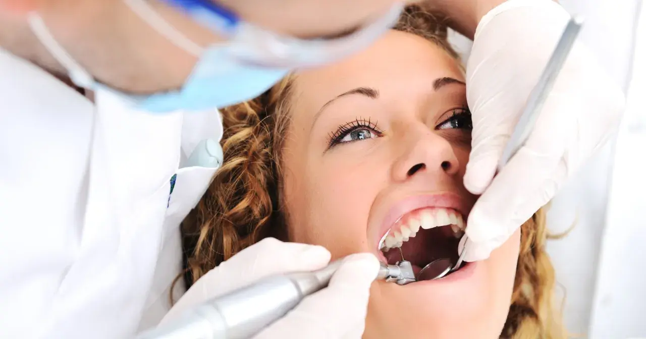 Kobieta u dentysty, zastanawia się ile kosztuje plomba prywatnie. Dentysta w rękawiczkach używa wiertła i lusterka.