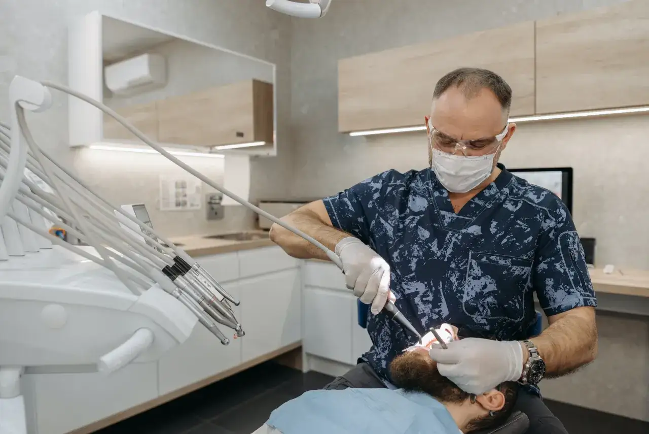Dentysta w masce i okularach wykonuje zabieg w sedacji. Pacjent leży na fotelu, a dentysta używa wiertła.