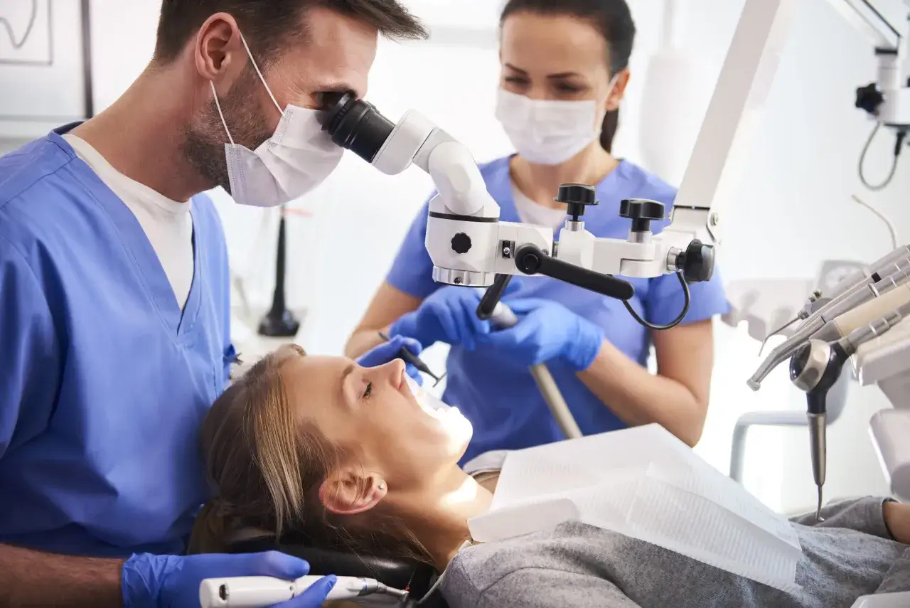 Dentysta z mikroskopem i asystentka wykonują zabieg. Pacjentka w gabinecie, gdzie można skorzystać z usług chirurgia stomatologiczna NFZ.