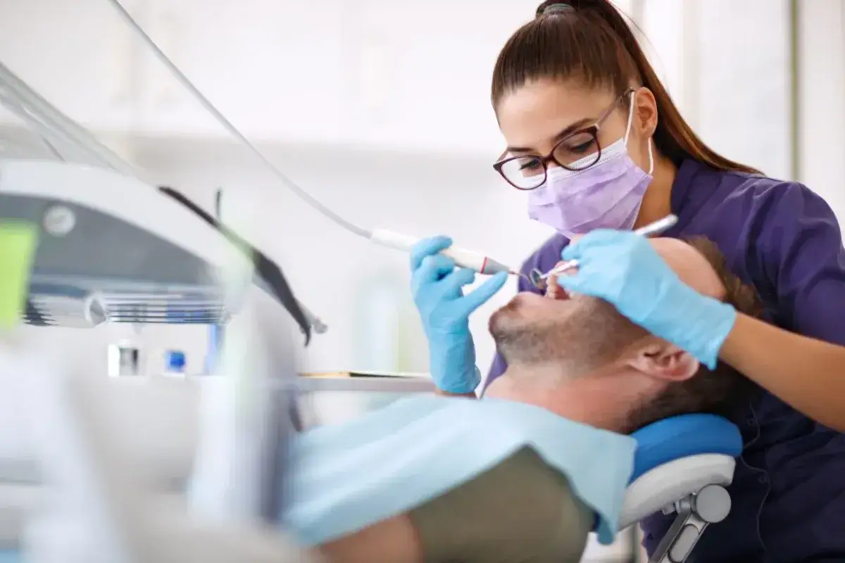 Dentystka w masce i rękawiczkach bada zęby pacjenta. Czy do dentysty potrzebne jest skierowanie? Zazwyczaj nie, wizyta jest możliwa po umówieniu terminu.