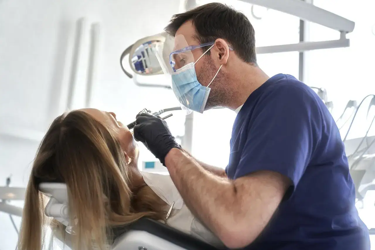 Dentysta w masce i przyłbicy bada pacjentkę. Zastanawiasz się, ile kosztuje usunięcie ósemki na NFZ?