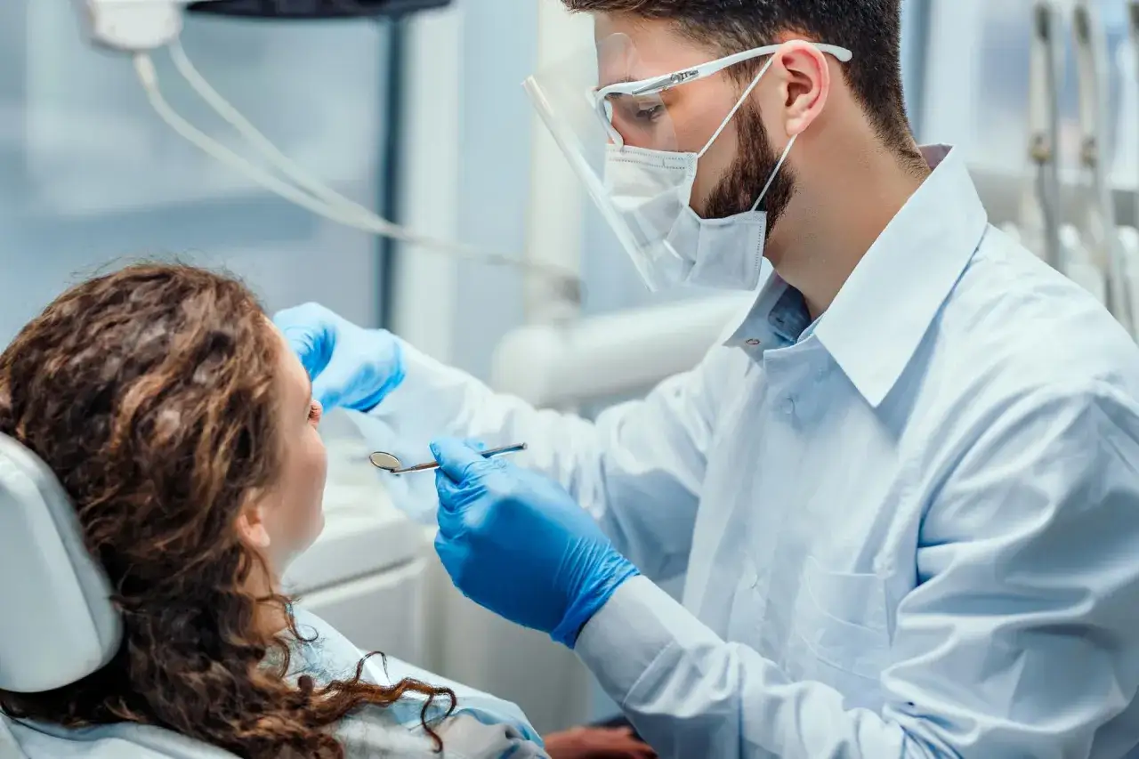 Dentysta w maseczce i przyłbicy wykonuje czyszczenie zębów na NFZ. Pacjentka siedzi na fotelu.
